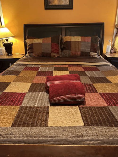 A neatly made bed with a patchwork quilt in warm colors and a folded red towel on top. Soft lighting in the background.