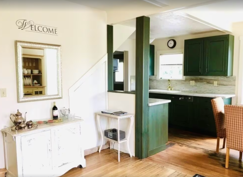 A cozy kitchen with green cabinets, wooden floors, and a welcoming decor featuring a mirror and a wine bottle.