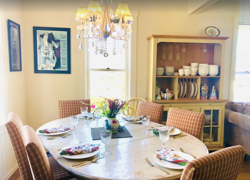 A cozy dining room with a round table set for a meal, featuring a chandelier and a sideboard with dishes.