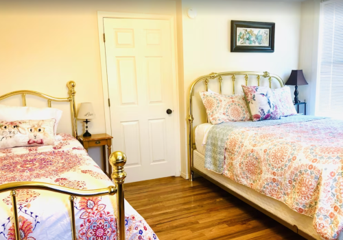 A bright bedroom featuring two gold metal beds with colorful patterned bedding and a small nightstand.
