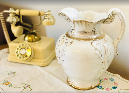 A vintage yellow telephone next to an ornate white vase with gold accents on a decorative tablecloth.