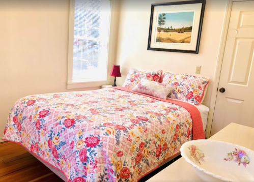 Cozy bedroom with a floral quilt, bedside lamp, and artwork on the wall, featuring a bowl on a dresser.