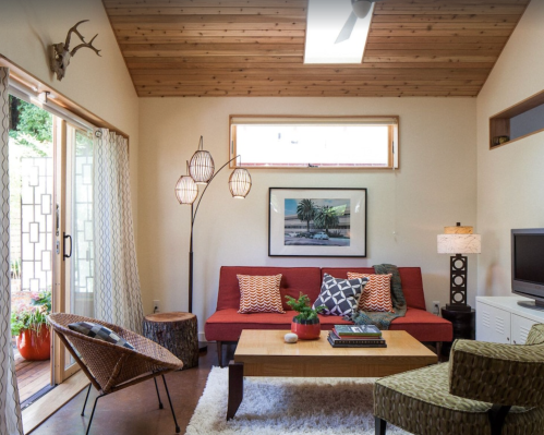 Cozy living room with a red sofa, wooden ceiling, decorative lamps, and a coffee table, featuring plants and artwork.