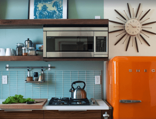 A modern kitchen featuring an orange Smeg fridge, stainless steel microwave, and a gas stove with fresh produce.