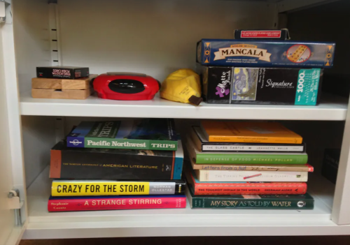 A shelf with books and games, including "Crazy for the Storm" and "Mancala," alongside various other titles and items.