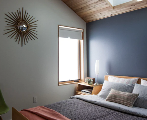 Cozy bedroom with a wooden bed, patterned bedding, a modern lamp, and a sunburst wall clock. Natural light from the window.