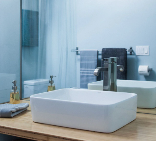 A modern bathroom sink with a square basin, faucet, and a mirror reflecting a towel and toilet in the background.