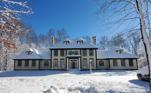 A large, elegant house covered in snow, surrounded by trees under a clear blue sky.