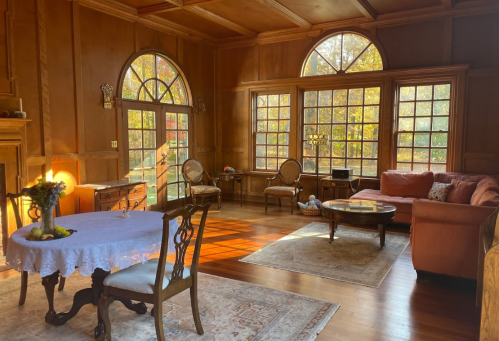 Cozy living room with wooden walls, large windows, a dining table, and comfortable seating, illuminated by warm sunlight.