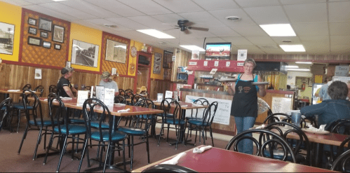 A diner interior with empty tables, a waitress holding a tray, and customers seated in the background.