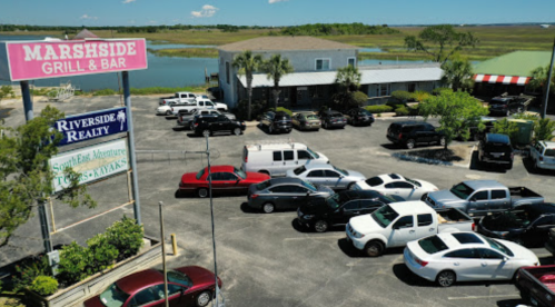 Aerial view of Marshside Grill & Bar with a busy parking lot and nearby water, surrounded by greenery.