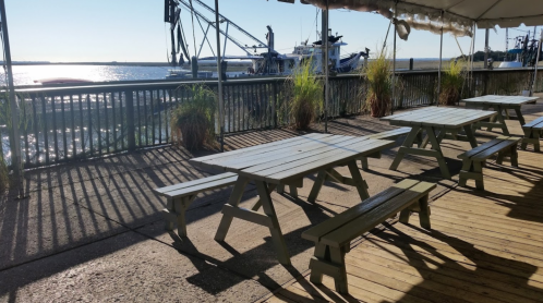 Outdoor seating area with wooden tables and benches overlooking a waterfront and boats in the background.