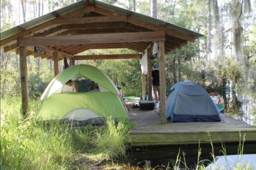Two tents on a wooden platform by a serene waterway, surrounded by lush greenery and trees.