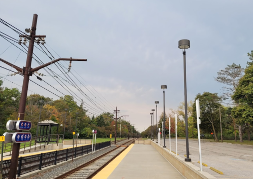A quiet train station platform with tracks, lights, and trees in the background under a cloudy sky.