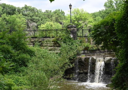 A serene scene of a small waterfall flowing under a stone bridge, surrounded by lush greenery and trees.