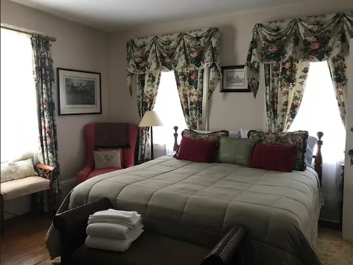 Cozy bedroom with a large bed, floral curtains, and a red armchair, featuring soft lighting and decorative pillows.