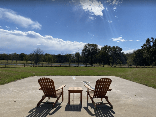 Two wooden chairs and a small table on a concrete patio, overlooking a grassy field and trees under a blue sky.