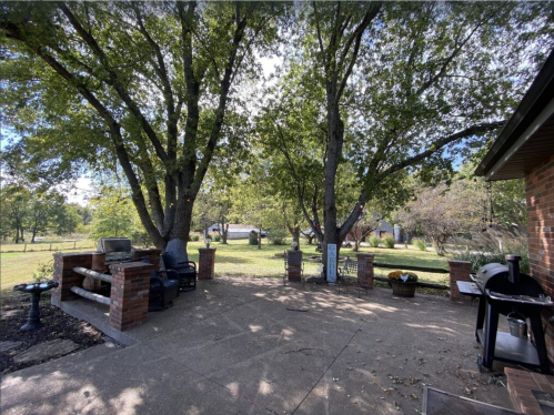 A spacious outdoor patio with two large trees, seating areas, and a grassy yard in the background under a clear sky.