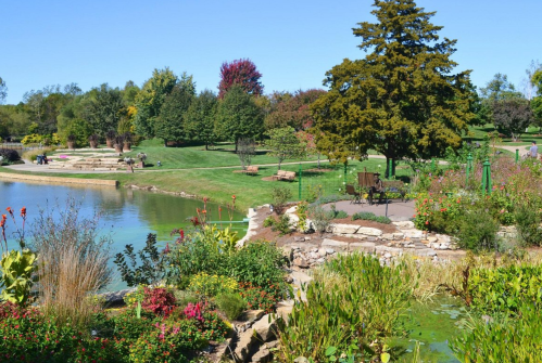 A serene park scene featuring a pond, lush greenery, colorful flowers, and benches under a clear blue sky.