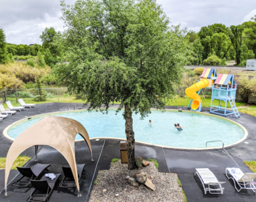 Aerial view of a pool with a slide, surrounded by trees, lounge chairs, and a shaded area. Two children are swimming.