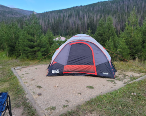 A gray and red tent set up on a gravel campsite surrounded by green trees and mountains in the background.