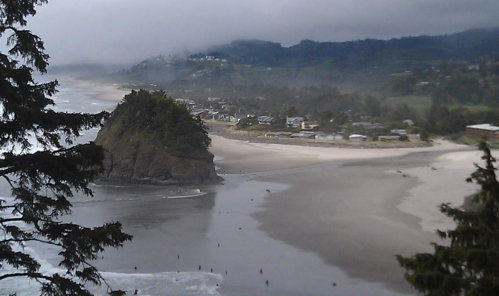 A scenic view of a beach with a rocky outcrop, surrounded by trees and misty hills in the background.