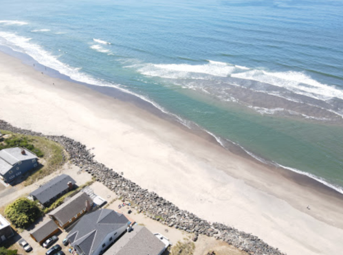 Aerial view of a sandy beach with gentle waves, bordered by houses and rocky shoreline under a clear blue sky.