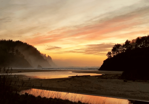 Sunset over a tranquil beach, with silhouettes of hills and trees reflected in the water. Soft clouds in the sky.