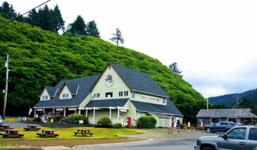 A green hillside surrounds a large, rustic building with a peaked roof and picnic tables in front.