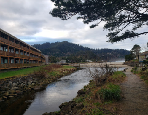 A serene landscape featuring a river, a wooden building, and mountains under a cloudy sky.