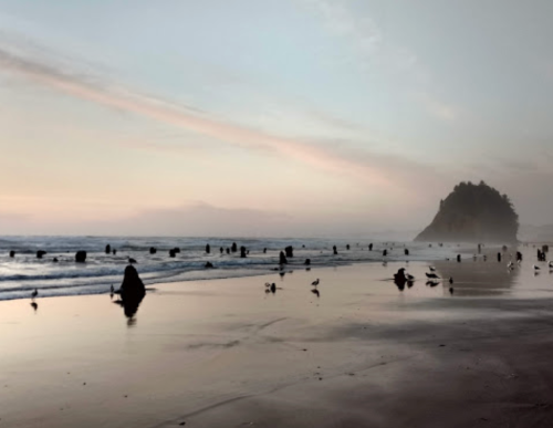 Silhouettes of people walking along a beach at sunset, with gentle waves and a rocky island in the background.