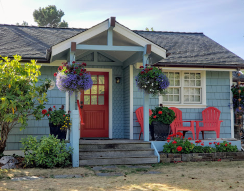 A charming blue cottage with a red door, flower baskets, and red chairs on the porch, surrounded by greenery.