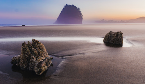 A serene beach scene at sunset, featuring two rocky formations and a distant island silhouette in the mist.