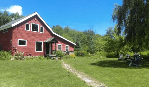 A red house with a sloped roof surrounded by green grass and trees under a clear blue sky.