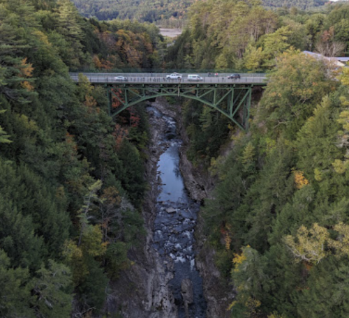 A green bridge spans a rocky river surrounded by dense trees and autumn foliage.