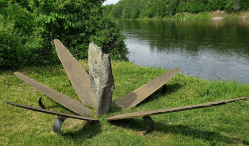 A unique sculpture resembling a flower, made of metal and stone, sits by a river surrounded by greenery.