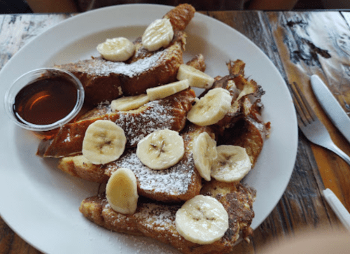 A plate of French toast topped with banana slices and powdered sugar, served with a side of syrup.