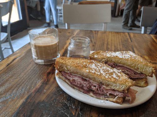 A plate with a sandwich filled with ham and a glass of iced coffee on a wooden table.