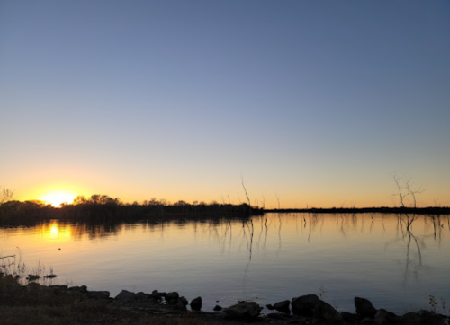 Sunset over a calm lake, with silhouettes of trees and rocks along the shore, creating a serene landscape.