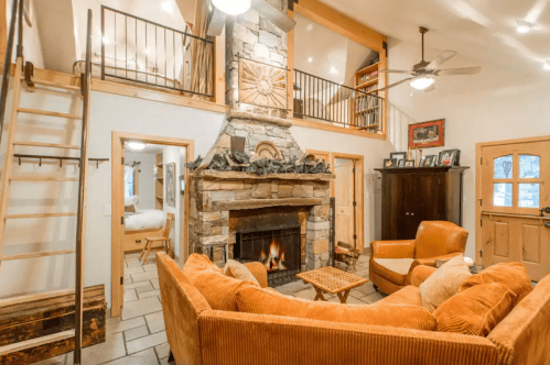 Cozy living room with a stone fireplace, orange sofas, and wooden accents, featuring a staircase and warm lighting.