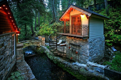 A cozy cabin with a balcony overlooks a stone bridge and a serene stream, surrounded by lush greenery.