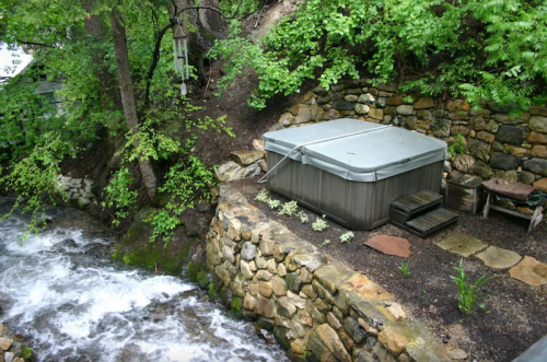 A hot tub sits beside a flowing stream, surrounded by lush greenery and stone landscaping.