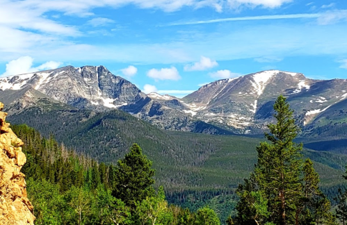 A scenic view of snow-capped mountains surrounded by lush green forests under a blue sky with scattered clouds.