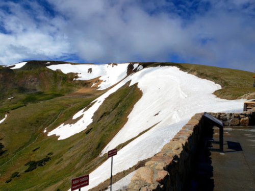 A scenic view of a snow-covered mountain slope with green grass and a stone wall in the foreground under a blue sky.