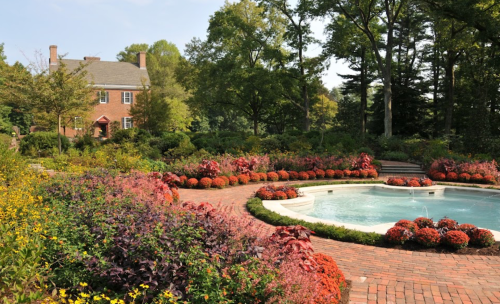 A landscaped garden with colorful flowers surrounding a circular pool and a brick pathway, leading to a brick house.