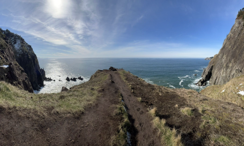 A panoramic view of a rugged coastline with cliffs, ocean waves, and a clear blue sky. A dirt path leads to the edge.
