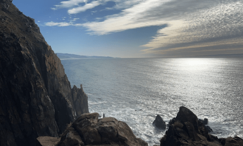 A scenic view of rocky cliffs overlooking a calm ocean under a partly cloudy sky.