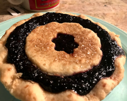 A round pie with a golden crust and a dark berry filling, featuring a decorative center and sparkling sugar on top.
