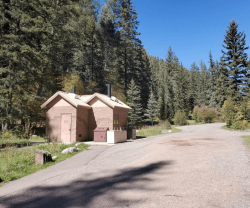 Two rustic restroom buildings beside a gravel path, surrounded by tall trees and clear blue skies.