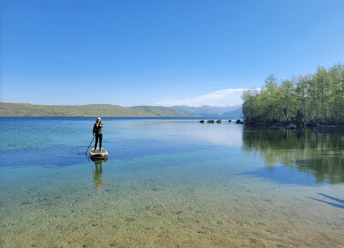 A person paddleboarding on a calm lake surrounded by trees and mountains under a clear blue sky.